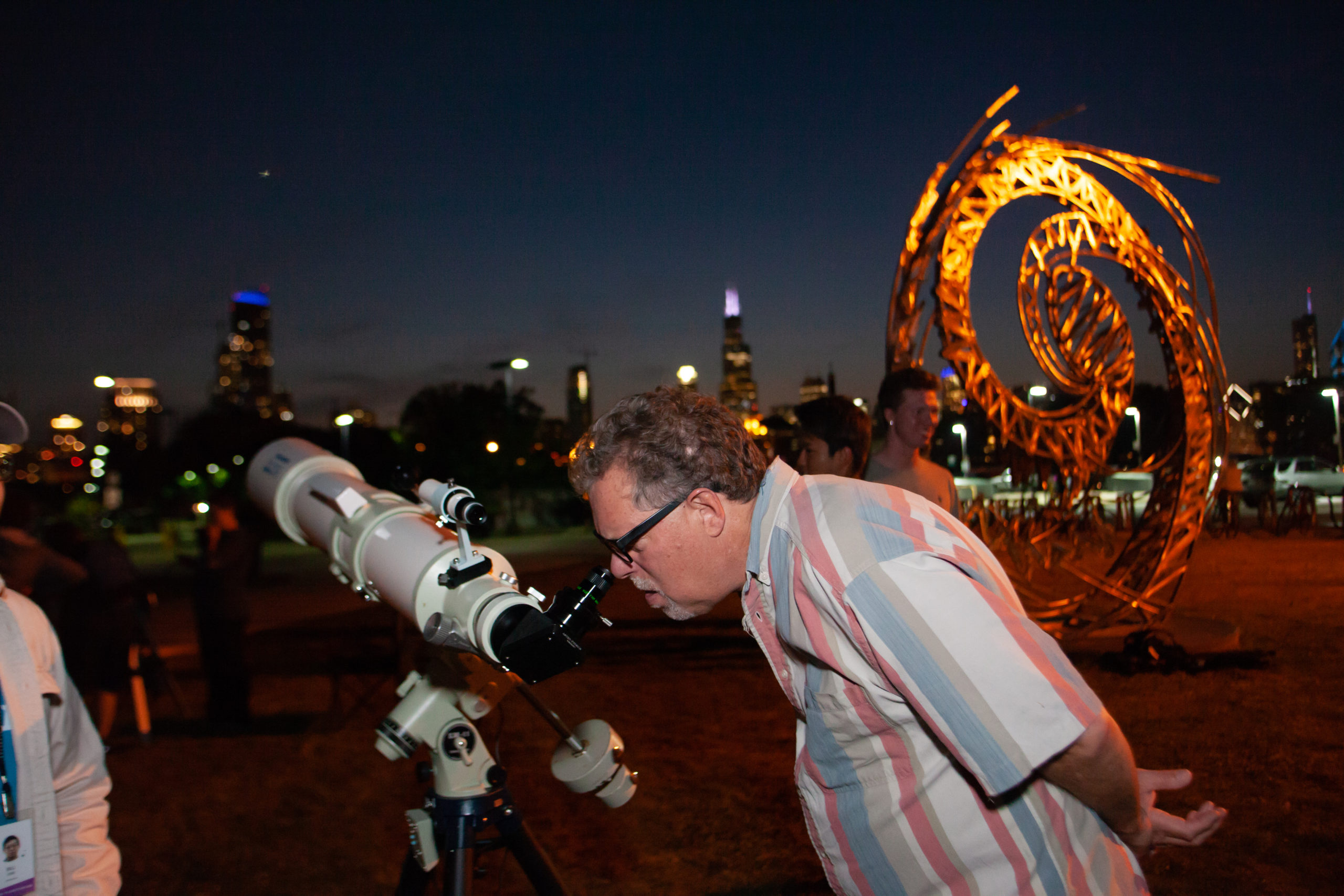 Individual looking through telescope in front of the Adler at night. Milky Way sculpture visible.