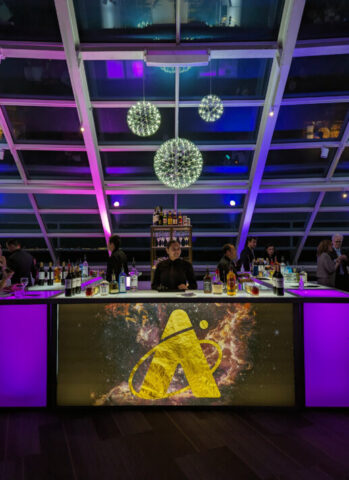 Central bar with prominent Adler monogram logo against a beautiful black and rust and gold colored JWST image. Purple backlit bar with three lit orbs hanging from the ceiling.