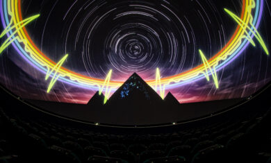 A theater with seats that has a 360 degree screen and projected onto the screen is an image of a dark night sky with white star trials, a rainbow heartbeat pulse, and three pyramids.