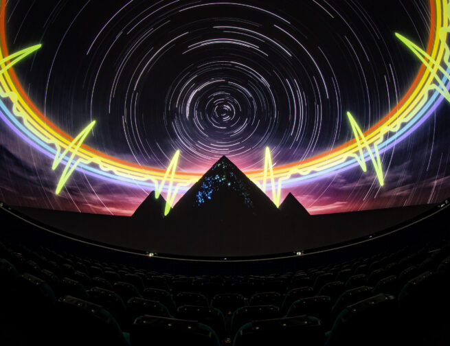 A theater with seats that has a 360 degree screen and projected onto the screen is an image of a dark night sky with white star trials, a rainbow heartbeat pulse, and three pyramids.