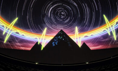 A theater with seats that has a 360 degree screen and projected onto the screen is an image of a dark night sky with white star trials, a rainbow heartbeat pulse, and three pyramids.