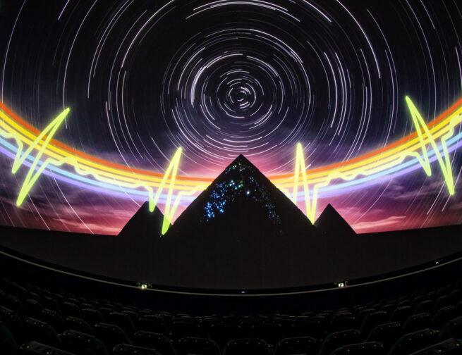 A theater with seats that has a 360 degree screen and projected onto the screen is an image of a dark night sky with white star trials, a rainbow heartbeat pulse, and three pyramids.