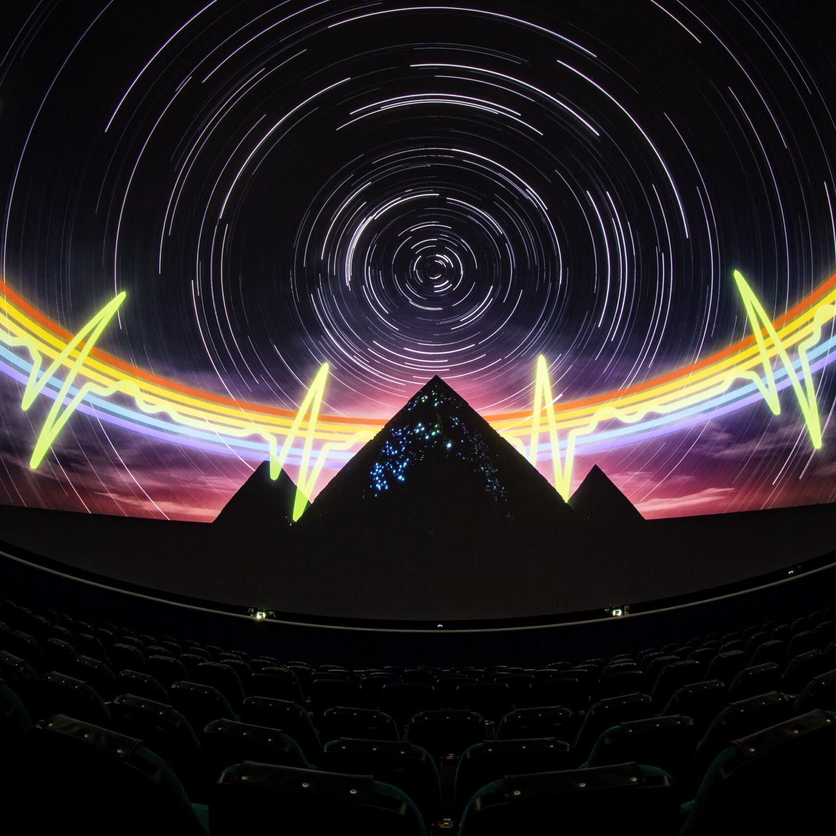 A theater with seats that has a 360 degree screen and projected onto the screen is an image of a dark night sky with white star trials, a rainbow heartbeat pulse, and three pyramids.