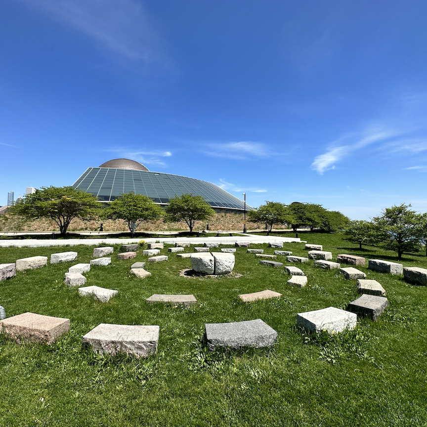 Rectangle rock blocks in a spiral pattern on green grass with trees and the Adler Planetarium in the background.