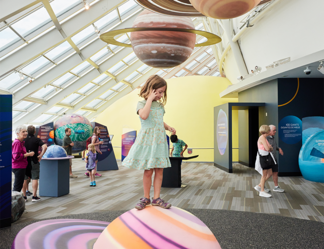 A child stands atop a pink, purple, and orange colored ringed planet in the Adler Planetarium’s exhibit, Other Worlds. Planet models hang from the ceiling above them while other guests interact with other features of the exhibit in the background.
