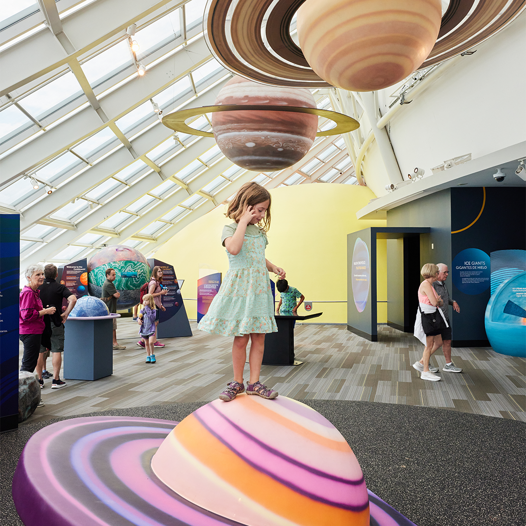 A child stands atop a pink, purple, and orange colored ringed planet in the Adler Planetarium’s exhibit, Other Worlds. Planet models hang from the ceiling above them while other guests interact with other features of the exhibit in the background.