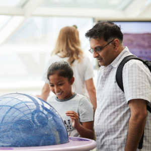 A child and an adult smile as they interact with a blue model that emulates the atmosphere of a gas giant planet in the Adler Planetarium’s exhibit, Other Worlds.