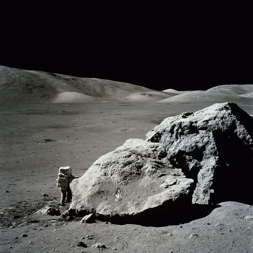 An astronaut in a space suit stands on the lunar surface next to a large lunar boulder.
