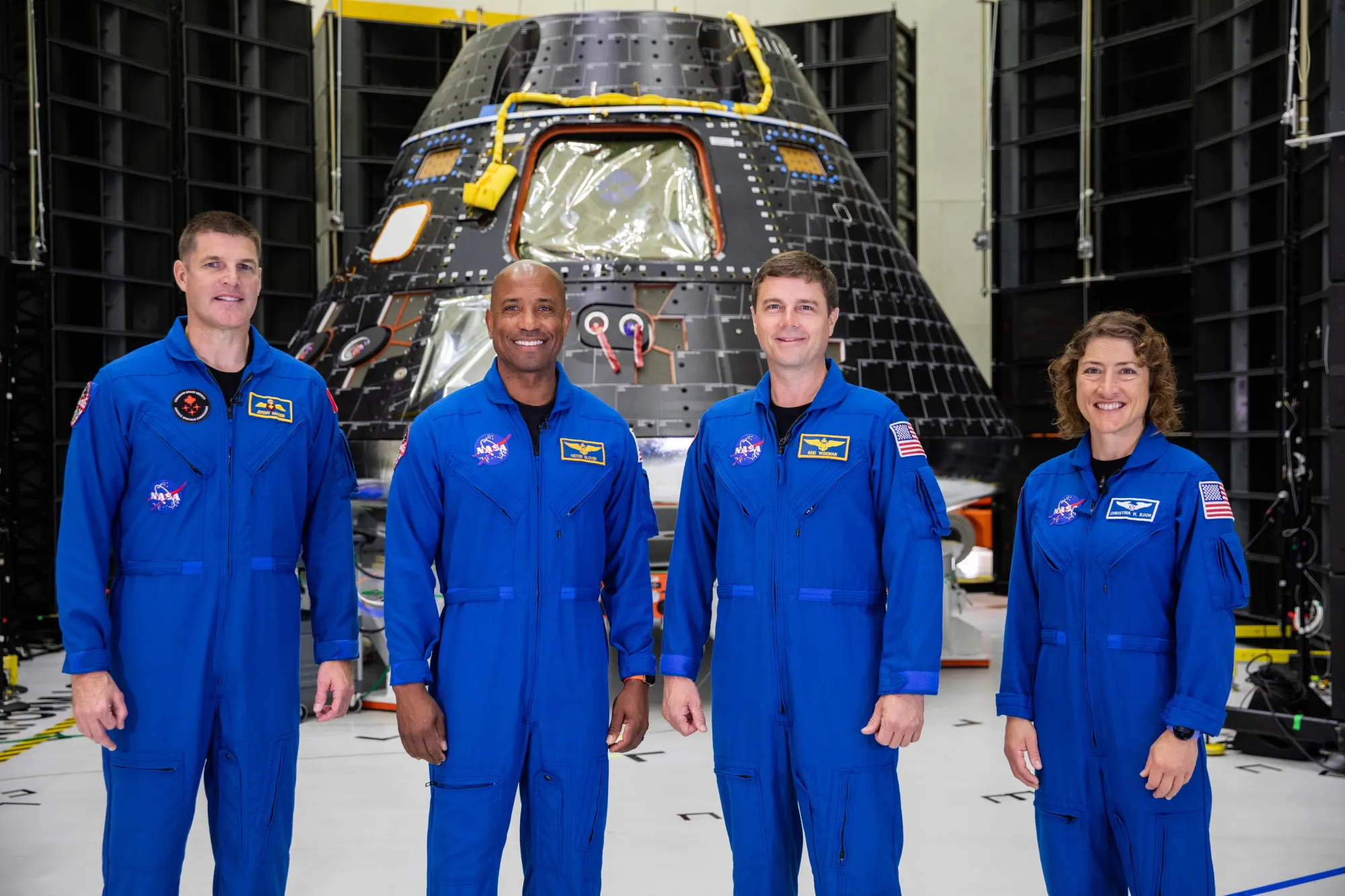Four people in bright blue jumpsuits pose smiling in front of a spacecraft. 