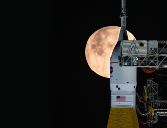 The tip of a rocket at the launch pad with a bright full Moon shining in the night sky behind it.