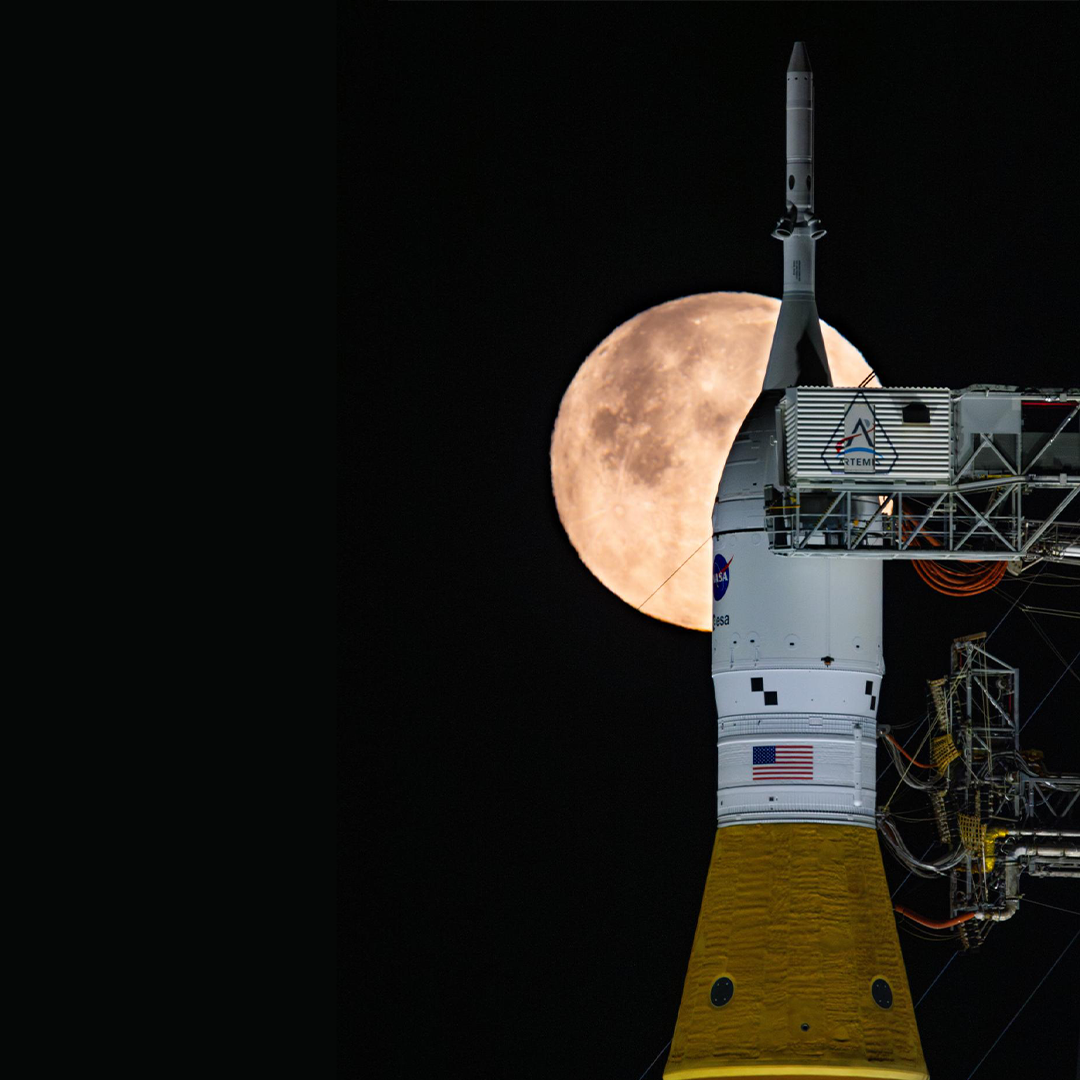 The tip of a rocket at the launch pad with a bright full Moon shining in the night sky behind it.
