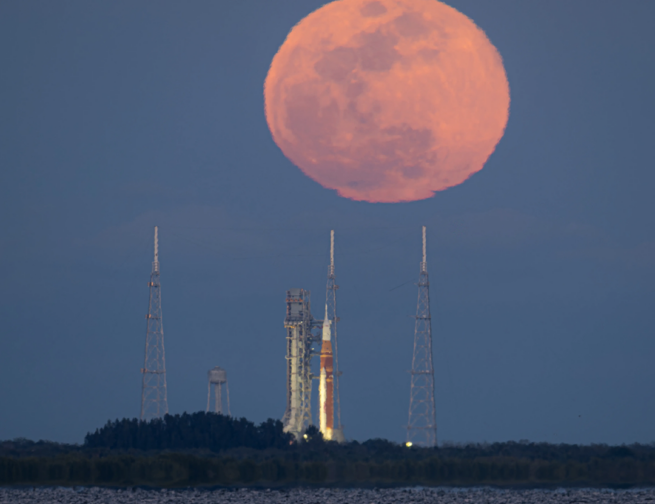 A bright orange full Moon rises behind a rocket on a launch pad at NASA’s Kennedy Space Center.
