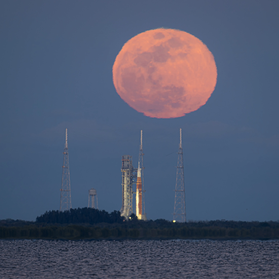 A bright orange full Moon rises behind a rocket on a launch pad at NASA’s Kennedy Space Center.