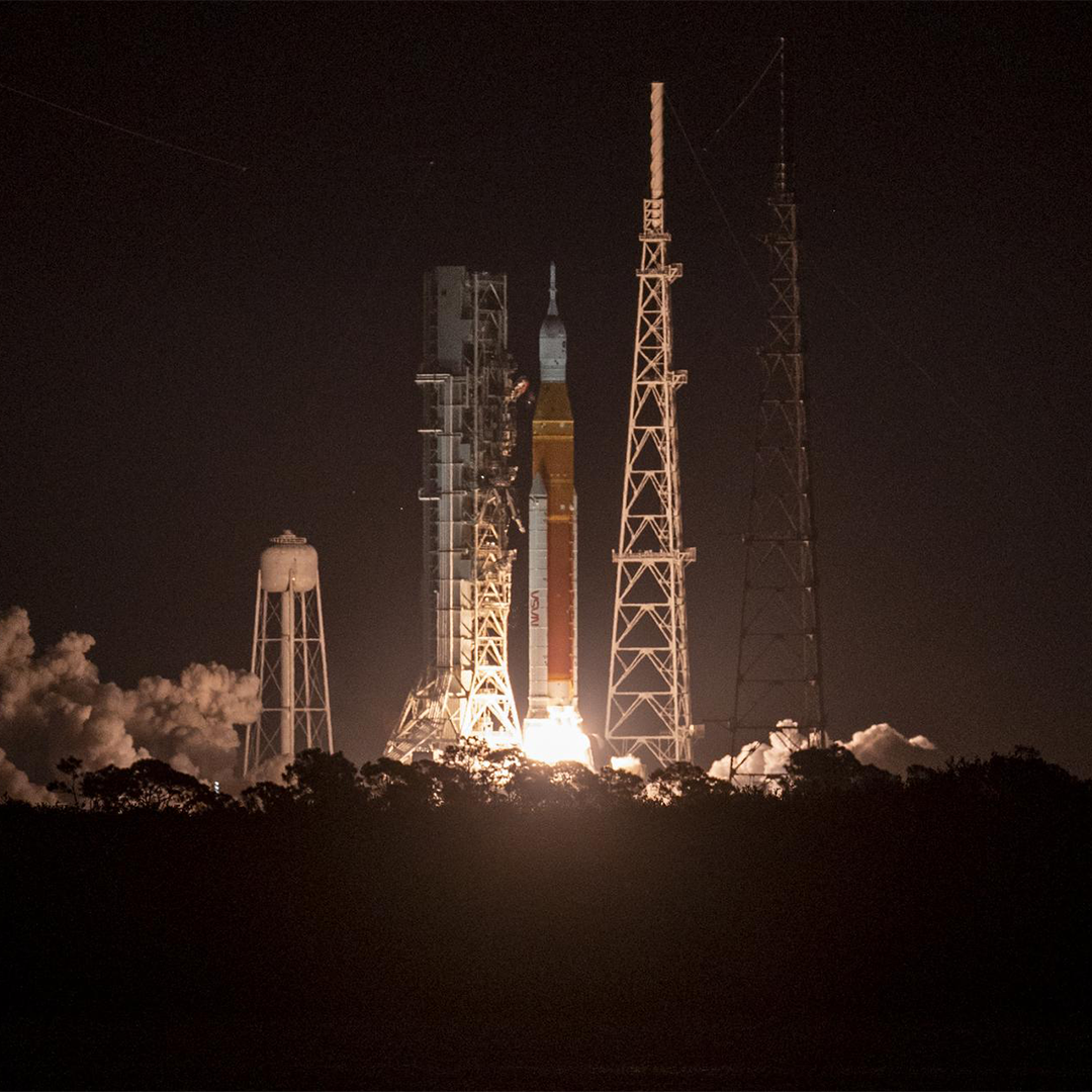 A rocket lifting off the launch pad, just seconds after launch. Fire and plumes of smoke appear at the base of the rocket as it begins to lift off. 