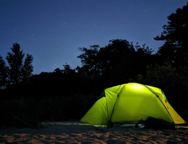 A green tent on sand with silhouetted trees and a blue night sky with stars