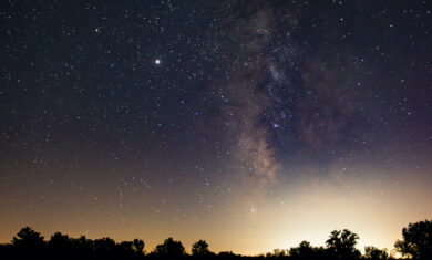 Milky Way in the sky and reflected in a lake with silhouetted trees between the sky and the lake taken at Middle Fork River Forest Preserve Image Credit: Nick Lake