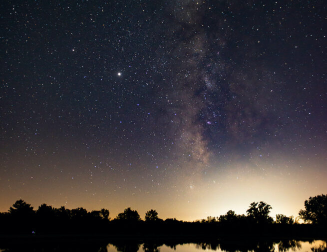 Milky Way in the sky and reflected in a lake with silhouetted trees between the sky and the lake taken at Middle Fork River Forest Preserve Image Credit: Nick Lake