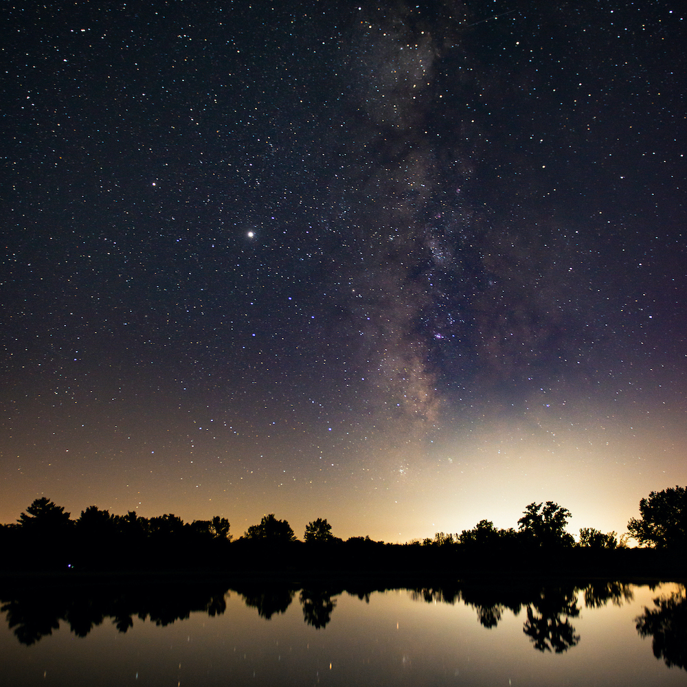 Milky Way in the sky reflected in a lake with silhouetted trees between the sky and the lake taken at Middle Fork River Forest Preserve Image Credit: Nick Lake