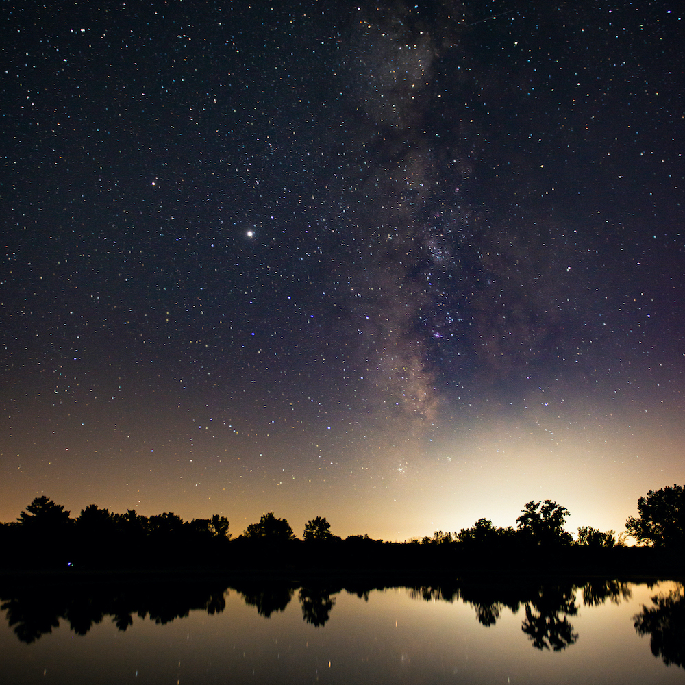 Milky Way in the sky and reflected in a lake with silhouetted trees between the sky and the lake taken at Middle Fork River Forest Preserve Image Credit: Nick Lake