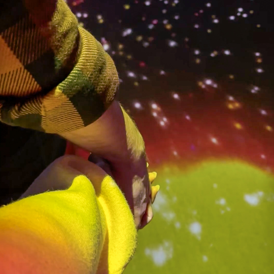 A close up of two people’s hands intertwined under colorful lights and star imagery at the Adler Planetarium