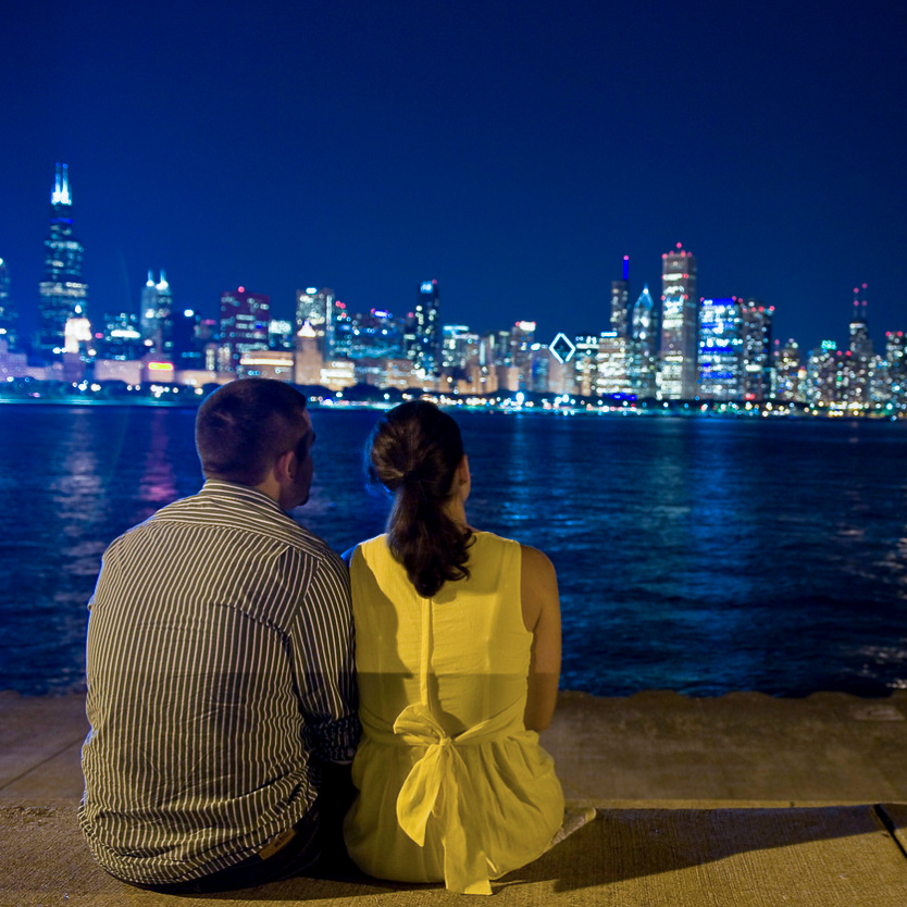 A couple sitting on the sidewalk outside the Adler Planetarium, enjoying the view of Lake Michigan and the Chicago skyline in the night sky.