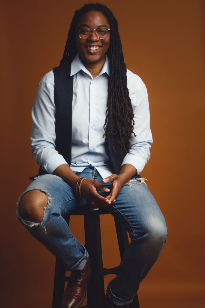 A photo of Dr. Christina Harrington sitting down on a stool while smiling at the camera.