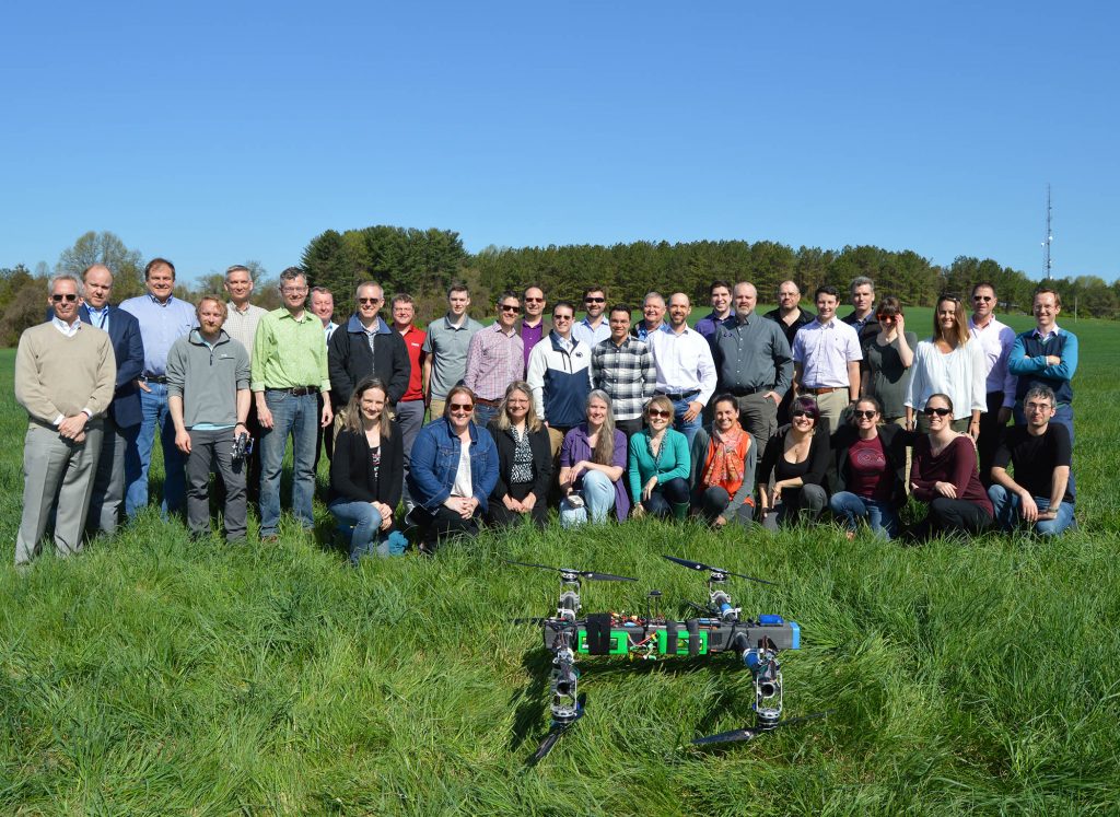 Dragonfly team in standing in two rows in a green field. in the foreground is the Dragonfly drone.