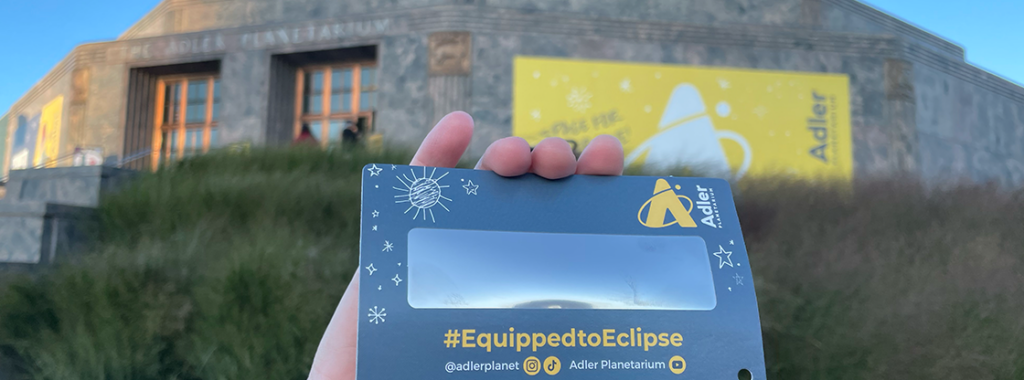 A solar eclipse viewer that says "#EquippedToEclipse" being held up by a hand in front of the Adler Planetarium
