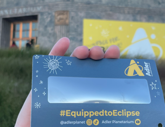 A solar eclipse viewer that says "#EquippedToEclipse" being held up by a hand in front of the Adler Planetarium