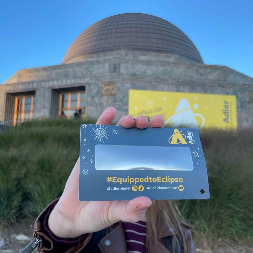 A solar eclipse viewer that says “#EquippedToEclipse” being held up by a hand in front of the Adler Planetarium