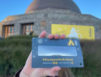 A solar eclipse viewer that says “#EquippedToEclipse” being held up by a hand in front of the Adler Planetarium.