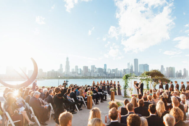 daytime wedding ceremony with a beautiful Chicago skyline view behind an ivy archway. Guests fill the seats on either side of the center aisle