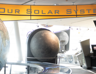 The entry way to Our Solar System exhibit. A large globe of Earth is to the right, the center is a model of the Moon and the words "Our Solar System" hangs above with grey letters and a bright orange background.