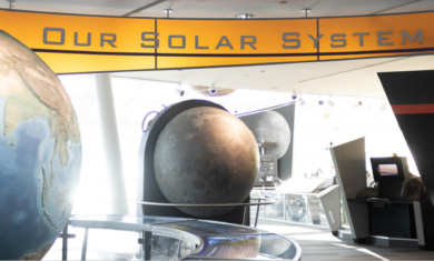 The entry way to Our Solar System exhibit. A large globe of Earth is to the right, the center is a model of the Moon and the words "Our Solar System" hangs above with grey letters and a bright orange background.