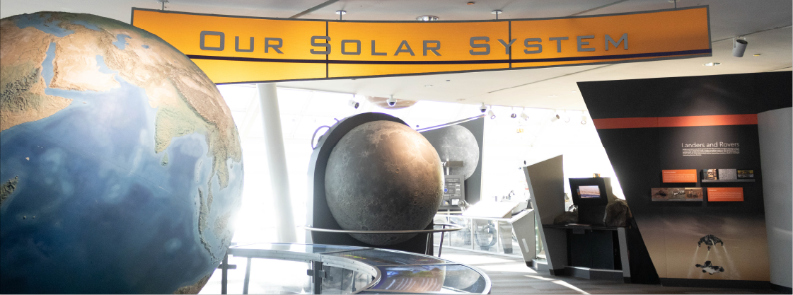 The entry way to Our Solar System exhibit. A large globe of Earth is to the right, the center is a model of the Moon and the words "Our Solar System" hangs above with grey letters and a bright orange background.