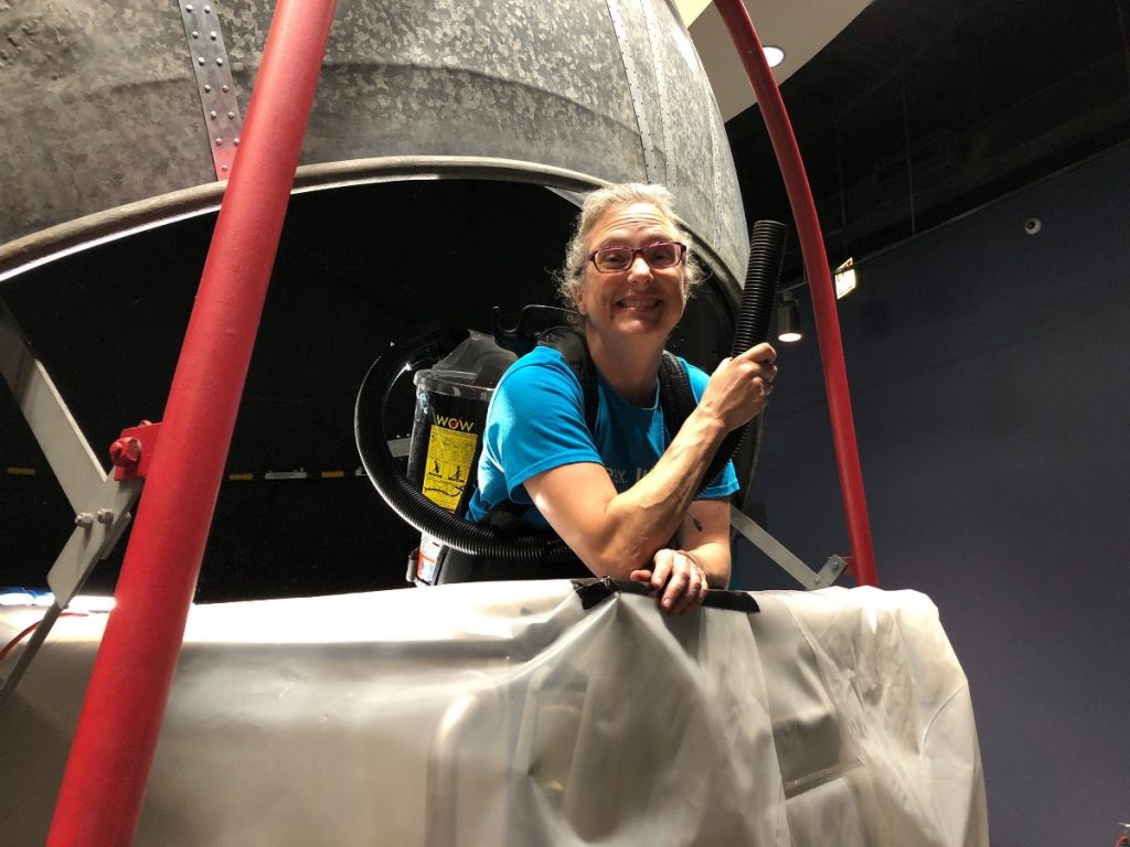 Michelle Nichols stands in the cart of the Atwood Sphere with a vacuum to collect dust as the sphere is painted.