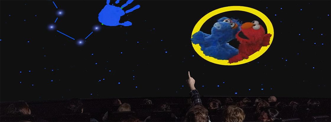 Dome theater showing Elmo and Hu Hu Zhu looking at stars and a blue handprint in the sky while a person from the audience sitting in a seat points up at the screen.