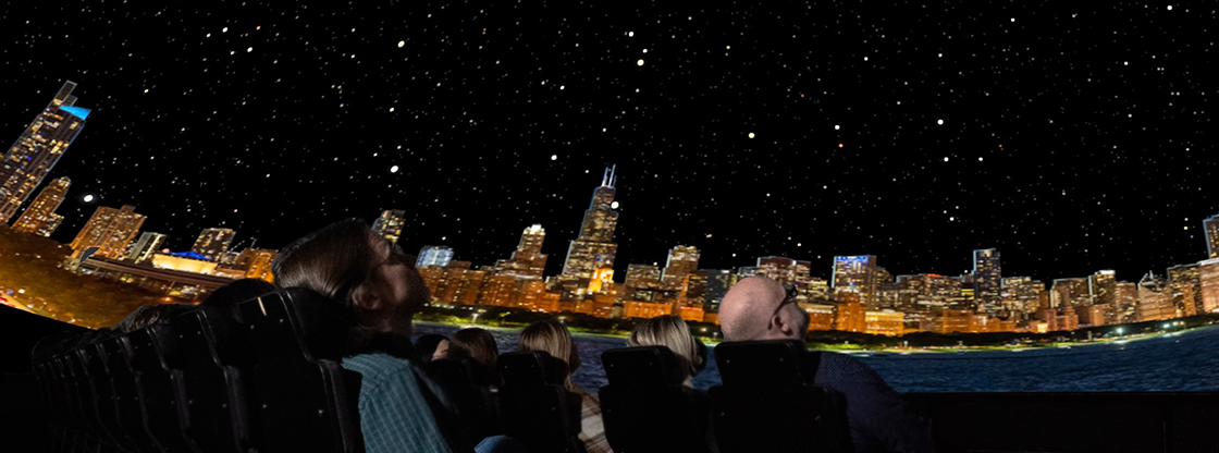 People sitting in seats looking up at a dome theater screen at a nighttime view of the Chicago skyline with hundreds of stars in the background behind the city