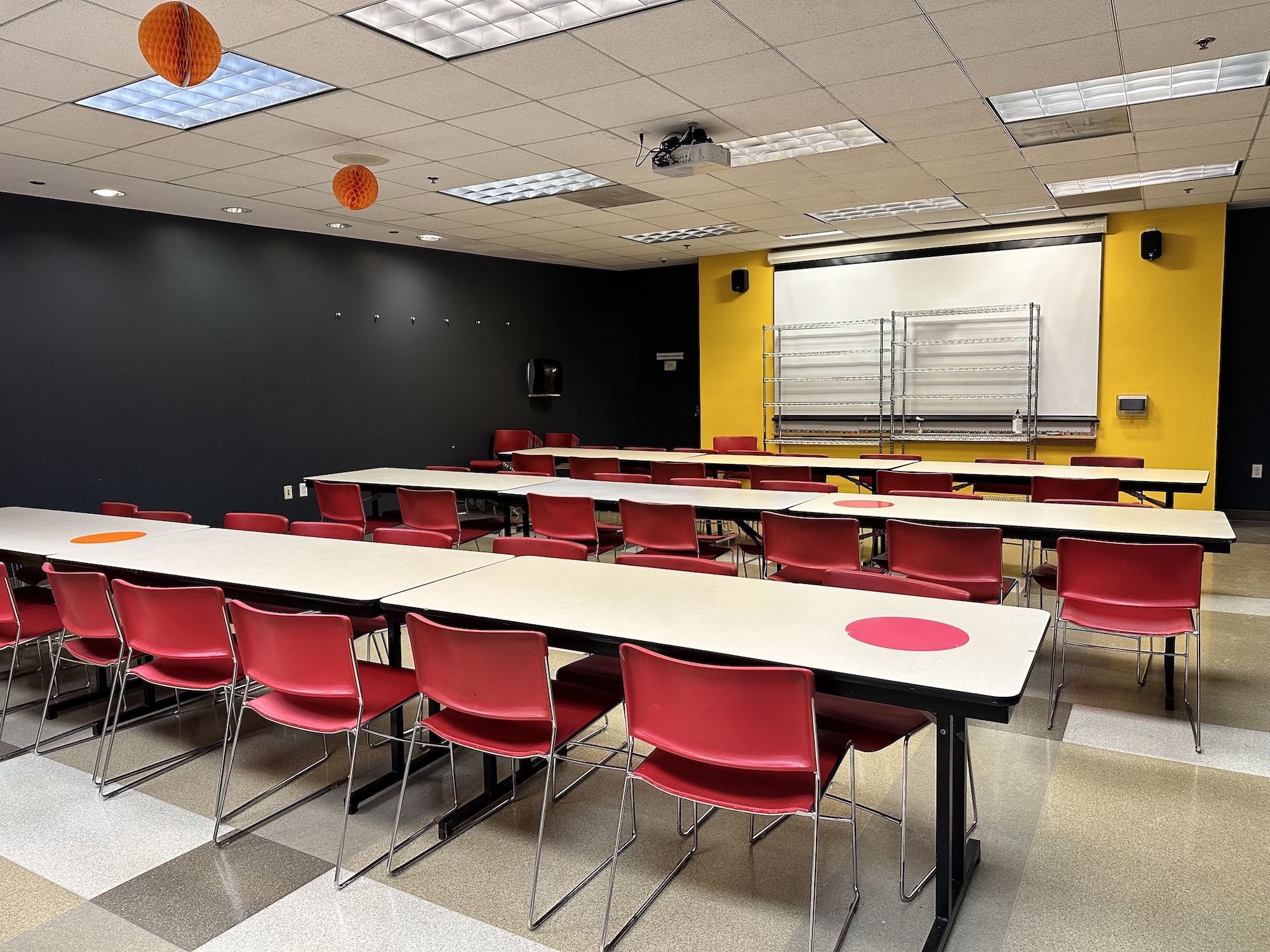 Interior image of Classroom A featuring long rectangular tables and red chairs lined up three in a row facing a yellow wall and whiteboard with some metal shelving in front of it