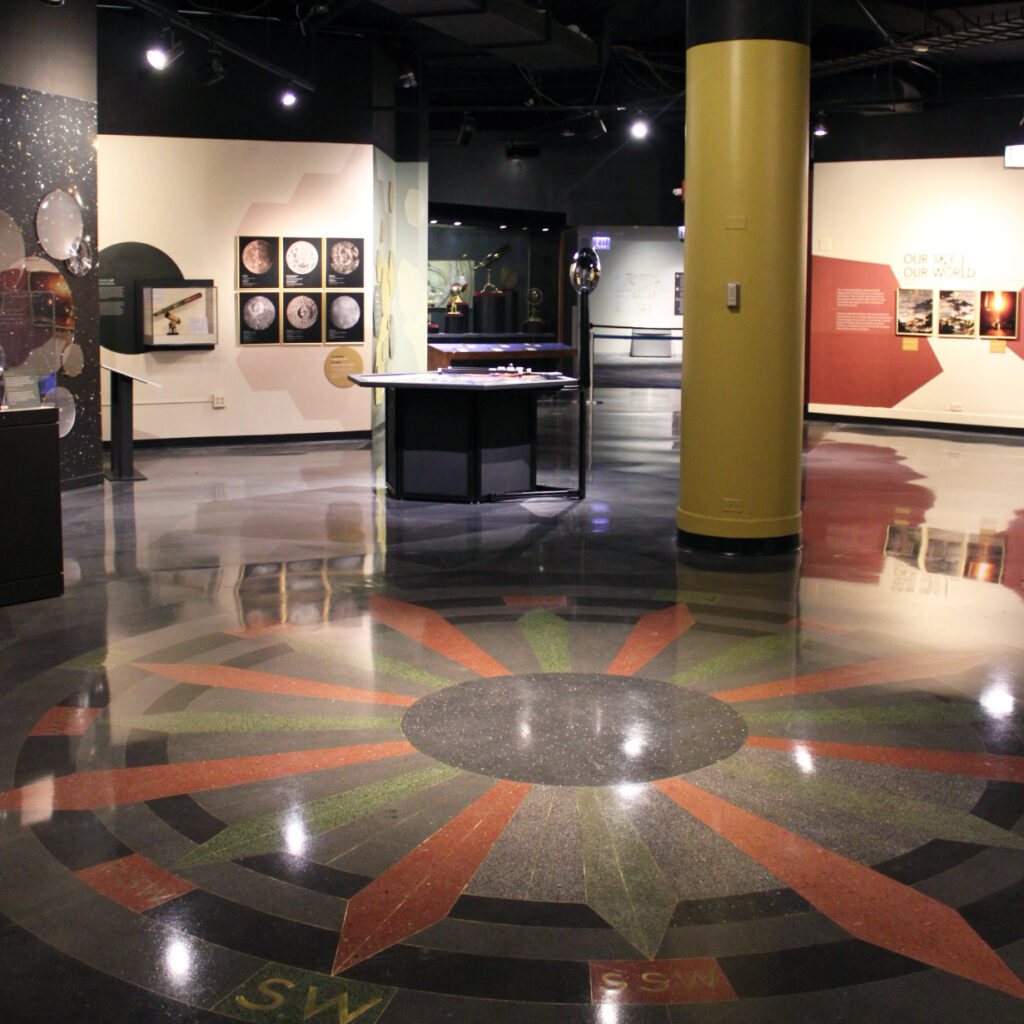 A room with a red, green, and black compass floor design that has a yellow column and hexagon table in the middle of the space, and pictures of the Moon, sunsets and large mirrors or lens on the surrounding walls.