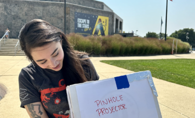 A person stands in front of the Adler Planetarium, holding a homemade pinhole projector to safely observe the Sun without solar viewers.
