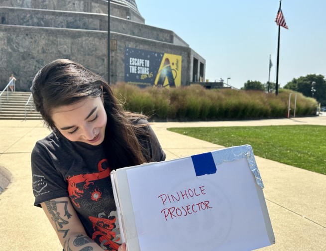 A person stands in front of the Adler Planetarium, holding a homemade pinhole projector to safely observe the Sun without solar viewers.