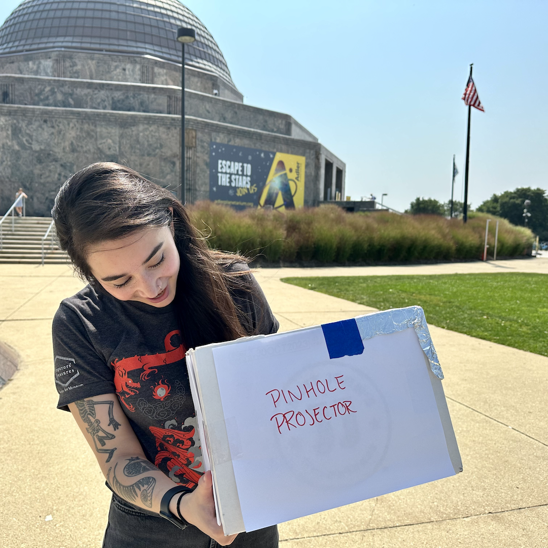 A person stands in front of the Adler Planetarium, holding a homemade pinhole projector to safely observe the Sun without solar viewers.