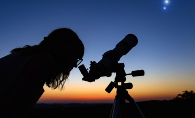 Person looking through telescope, observing two bright objects in the sky at dusk.