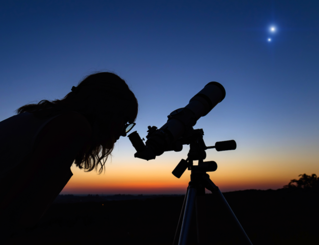 Person looking through telescope, observing two bright objects in the sky at dusk.