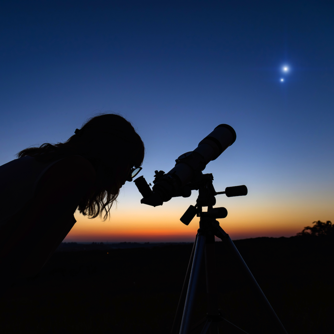 Person looking through telescope, observing two bright objects in the sky at dusk.