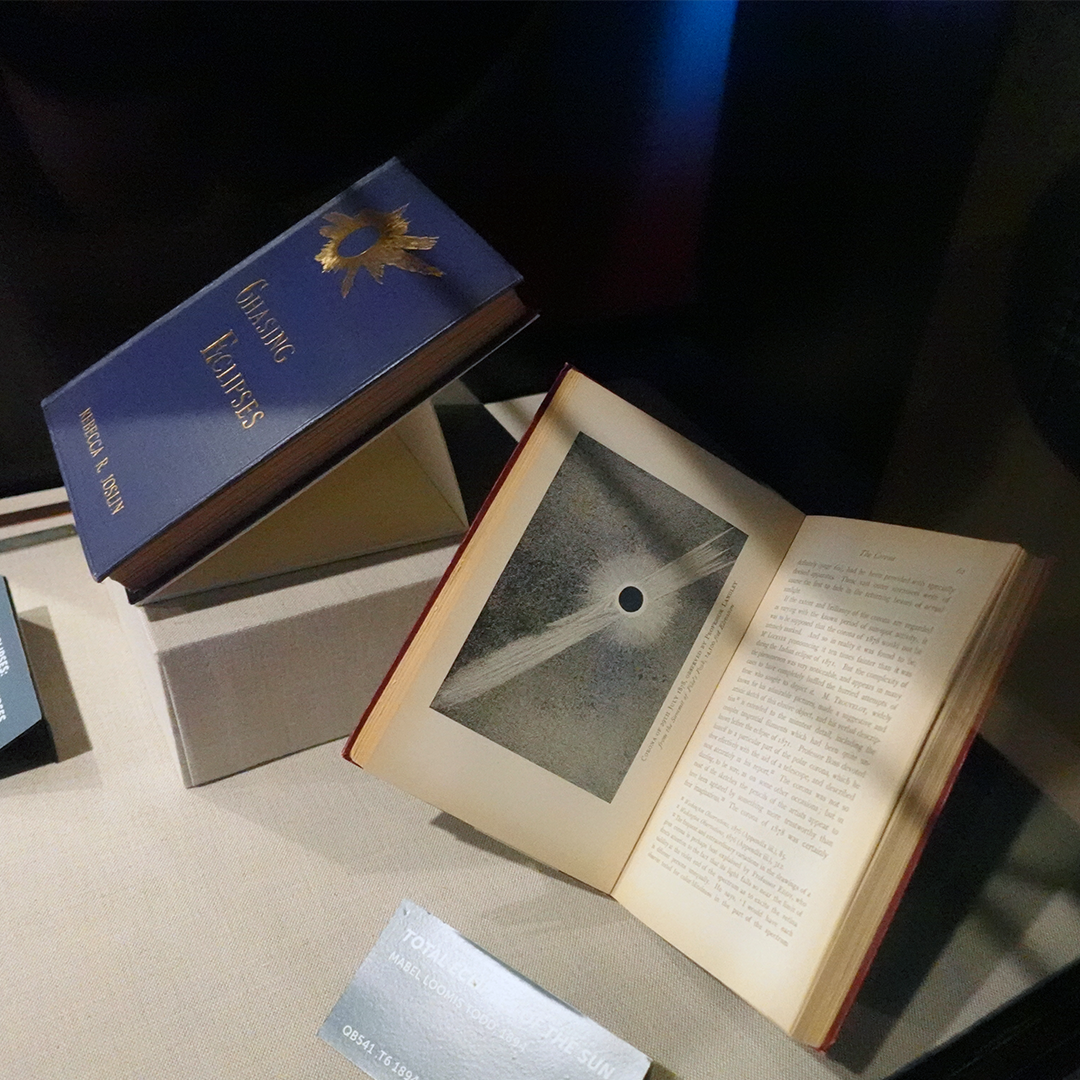 Two books on display in the exhibit Chasing Eclipses at the Adler Planetarium in Chicago, IL. The blue cover of Rebecca R. Joslin’s book Chasing Eclipses is seen on the right, next to an open copy of Total Eclipses Of The Sun by Mabel Loomis Todd, featuring a black and white illustration of a total solar eclipse. 