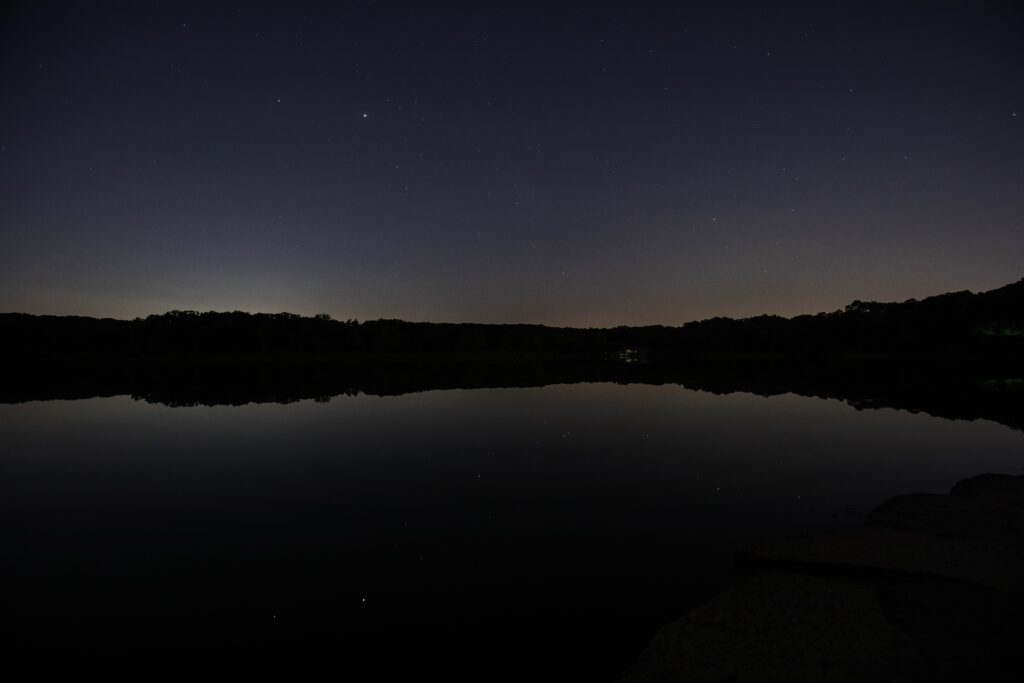 Dark blue night sky with stars reflected in a lake with silhouetted trees between the sky and lake at Maple Lake in the Forest Preserve of Cook County's Palos Preserves. Image Credit: Joe Occhiuzzo
