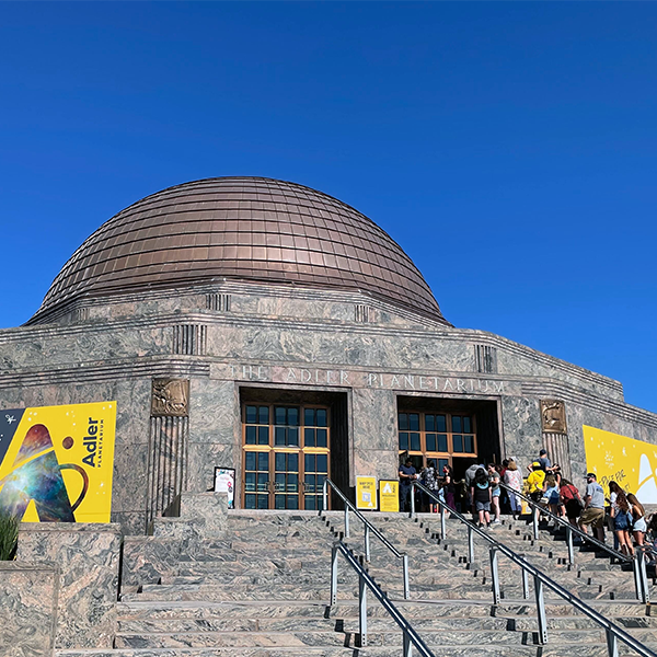 The Adler Planetarium on a sunny, summer day, with a line of guests on the museum steps. A bright blue sky contrasts the museum's yellow banners.