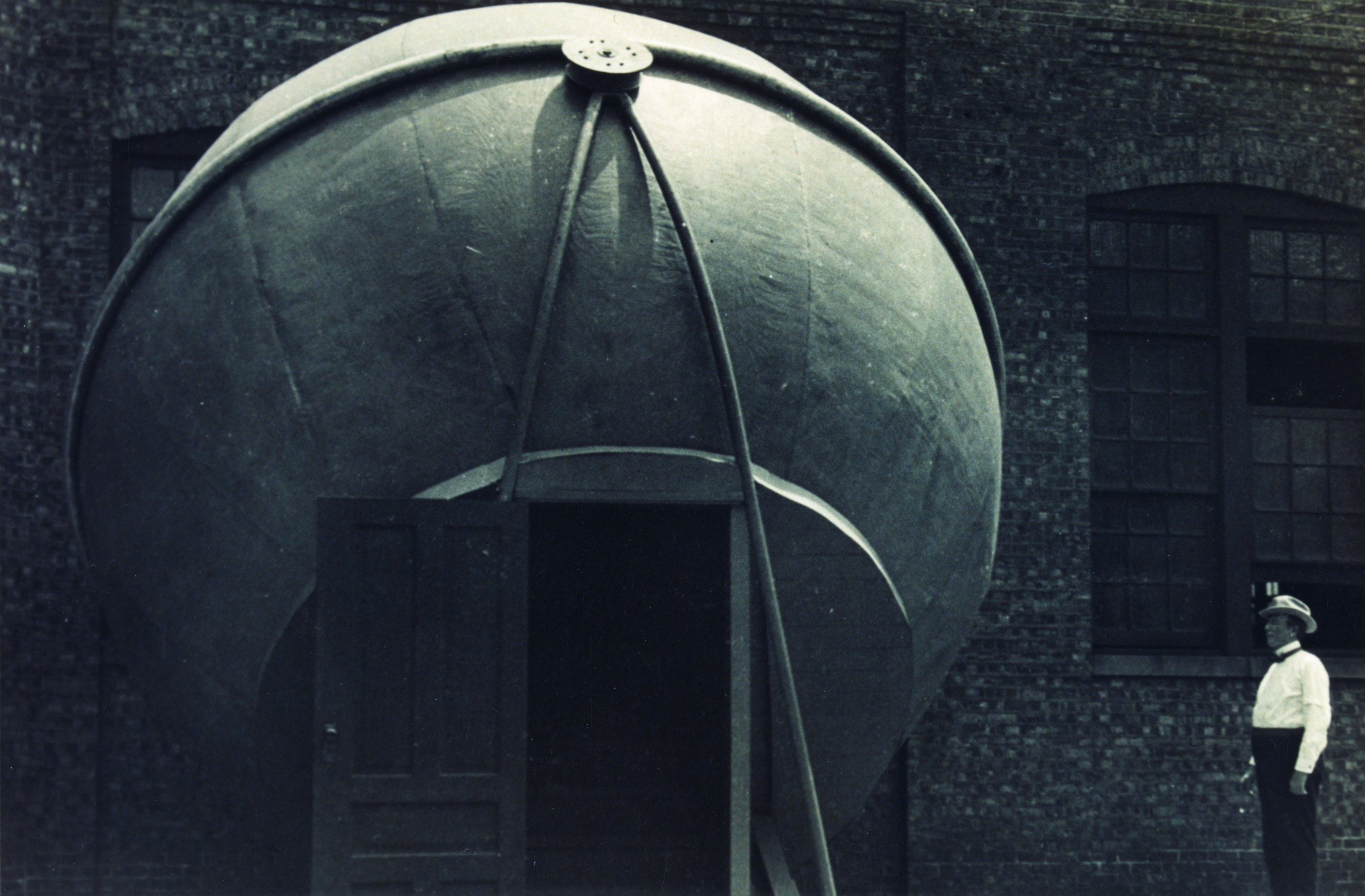  Image Caption: Outside a brick building the Atwood Sphere stands assembled with an open wooden door at the bottom. A man is standing to the right. c. 1913 Image Credit: Adler Planetarium Archives